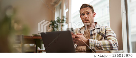 Portrait of cafe visitor sitting with laptop and mobile phone, looking confused, puzzled at camera 126013970