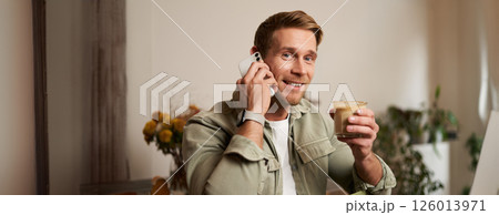 Image of smiling, good-looking young blond man with phone, drinking coffee in cafe, talking to someone over the telephone, answering a call Image of smiling, good-looking young blond man with phone, drinking coffee in cafe, talking to someone over the telephone, answering a call 126013971