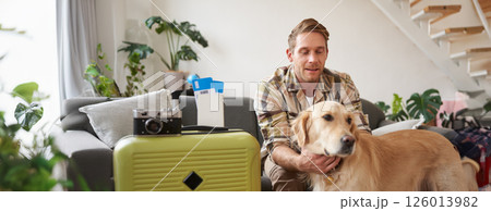 Portrait of young man, traveller, pets his dog, sits with suitcase and tickets, goes on holiday Portrait of young man, traveller, pets his dog, sits with suitcase and tickets, goes on holiday 126013982