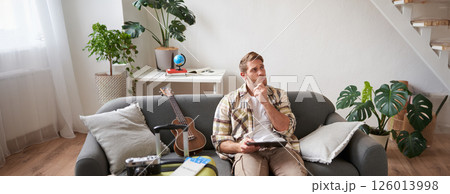 Image of young man with tablet, sitting in living room with suitcase, reading, studying the route, preparing for a vacation 126013998