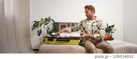 Portrait of young man packing his luggage for a trip, going on holiday, sitting on bed and putting clothes in suitcase, preparing for his flight Portrait of young man packing his luggage for a trip, going on holiday, sitting on bed and putting clothes in suitcase, preparing for his flight 126014032
