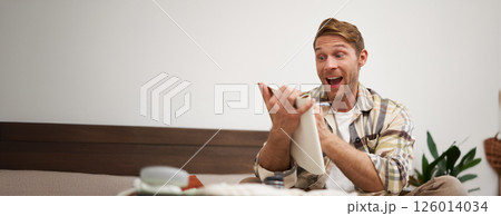 Portrait of charismatic, handsome man, tourist looking at notebook, getting ready for journey, packing suitcase, checking list of items on journal 126014034
