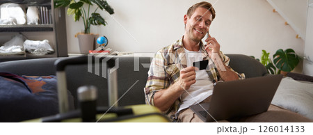 Portrait of smiling guy, tourist booking hotel, confirming purchase over the phone, holding credit card and laptop Portrait of smiling guy, tourist booking hotel, confirming purchase over the phone, holding credit card and laptop 126014133