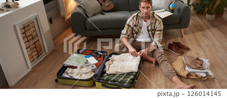 Travelling and tourism concept. Happy young man, sits on floor with luggage opened, packing suitcase for holiday, going on vacation, putting clothes inside a bag Travelling and tourism concept. Happy young man, sits on floor with luggage opened, packing suitcase for holiday, going on vacation, putting clothes inside a bag 126014145