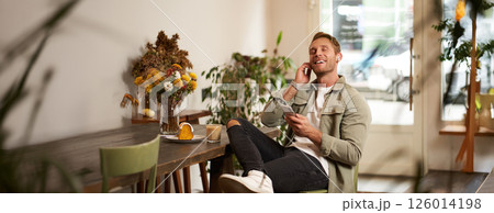 Portrait of handsome happy young man, relaxing in a coffee shop, spending time in cafe, listening to music in wireless headphones, holding smartphone, putting on his favourite song 126014198