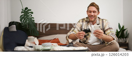 Smiling tourist, man with suitcase, sits on bed, pays online, books a hotel, holds credit card and mobile phone, uses an app to purchase in internet 126014203