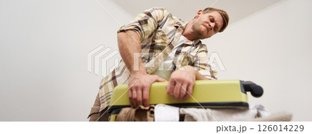 Image of young man, tourist struggling to close his suitcase full of clothes, pushing on bag, trying to press items inside luggage to shut the zipper, packing for travelling abroad, going on holiday 126014229