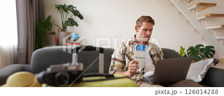 Angry man holding his flight tickets, staring upset at laptop screen, having problems with his holiday tour Angry man holding his flight tickets, staring upset at laptop screen, having problems with his holiday tour 126014288