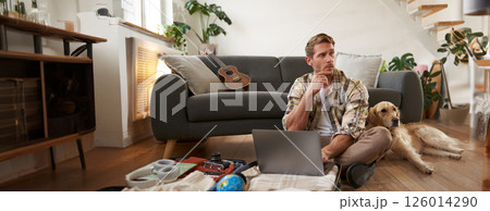 Portrait of young man, traveller going on holiday with a dog, packing his suitcase, sitting with laptop on his luggage, thinking, making online booking 126014290