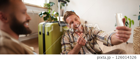 Smiling young man with suitcase, takes selfies on smartphone, blogger posts about his holiday tour on social media, using mobile phone Smiling young man with suitcase, takes selfies on smartphone, blogger posts about his holiday tour on social media, using mobile phone 126014326