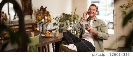 Charismatic, smiling young man, sitting and relaxing in cafe with cup of coffee, listens to music or podcast on wireless earphones, holds smartphone Charismatic, smiling young man, sitting and relaxing in cafe with cup of coffee, listens to music or podcast on wireless earphones, holds smartphone 126014333