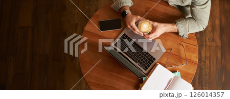 Top view. Male hands holding cup of coffee, man sitting at round table in a cafe, working on laptop, had his smartphone and notebook, drinking cappuccino Top view. Male hands holding cup of coffee, man sitting at round table in a cafe, working on laptop, had his smartphone and notebook, drinking cappuccino 126014357