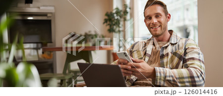 Portrait of young good-looking man, cafe visitor, sitting with laptop and smartphone, smiling at camera, freelancer working remotely from co-working space 126014416