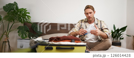 Portrait of handsome man, tourist, looking at his holiday tickets, booked vacation with travel agency, sitting on bed and packing clothes in suitcase 126014428