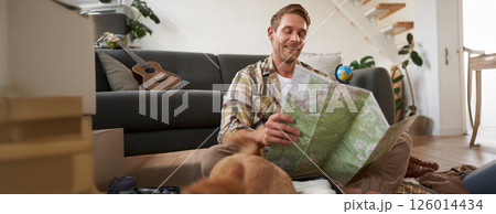 Portrait of young man, traveller, sitting packing suitcase, looking at travel map, choosing holiday destination, studying the route on vacation Portrait of young man, traveller, sitting packing suitcase, looking at travel map, choosing holiday destination, studying the route on vacation 126014434