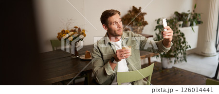 Portrait of young handsome man takes slefie on his mobile phone app, sits in cafe with glass of coffee, posting on social media application 126014441