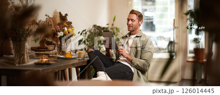 Portrait of young man sitting in cafe, spending time alone in coffee shop, reading on digital tablet, watching video, listening music in wireless headphones. Lifestyle and people concept Portrait of young man sitting in cafe, spending time alone in coffee shop, reading on digital tablet, watching video, listening music in wireless headphones. Lifestyle and people concept 126014445