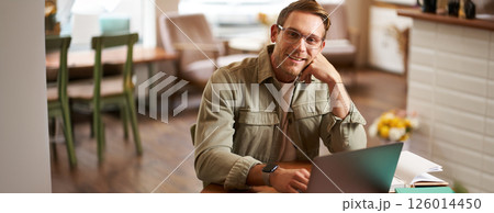 Remote work and freelance concept. Handsome man in glasses sits with notebook and laptop, studying online, working remotely from coffee shop, sitting in cafe with computer Remote work and freelance concept. Handsome man in glasses sits with notebook and laptop, studying online, working remotely from coffee shop, sitting in cafe with computer 126014450