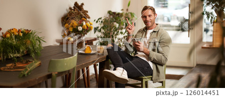 Portrait of happy handsome young man, sitting in cafe with mobile phone, wearing wireless headphones, listening to music or podcast while relaxing in coffee shop 126014451