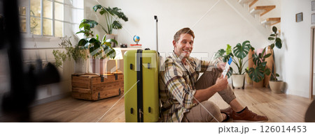 Portrait of handsome young man, tourist sitting with his suitcase, holding passport and flight tickets, going on holiday Portrait of handsome young man, tourist sitting with his suitcase, holding passport and flight tickets, going on holiday 126014453