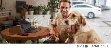 Close up portrait of smiling handsome european man with his dog in a cafe. Guy pets his golden retriever while working outdoors in coffee shop 126014504