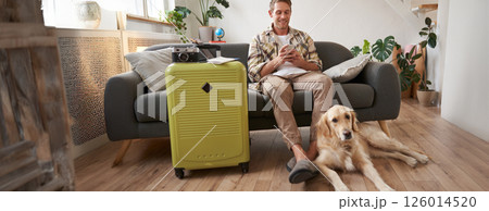 Portrait of guy with a dog, has packed suitcase, camera for going abroad and shooting pictures, dog resting near his legs, man looks at mobile phone and chats online Portrait of guy with a dog, has packed suitcase, camera for going abroad and shooting pictures, dog resting near his legs, man looks at mobile phone and chats online 126014520