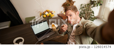 Portrait of serious man takes selfie with fist and threatening face, sits in cafe, poses in coffee shop in front of table with laptop and glass of coffee Portrait of serious man takes selfie with fist and threatening face, sits in cafe, poses in coffee shop in front of table with laptop and glass of coffee 126014574