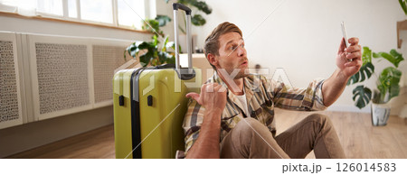 Portrait of happy tourist, man takes selfies before his flight, photos with suitcase, uses smartphone to post pictures on social media 126014583