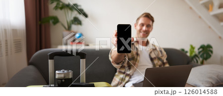 Portrait of handsome young man, showing smartphone screen, recommending mobile app for travelling or booking online, sitting with laptop and suitcase 126014588