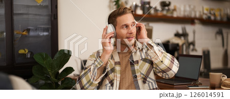 Lifestyle portrait of happy, excited young man, sitting in coffee shop with laptop and headphones, dancing on his chair and enjoying great quality sound, streaming favourite songs 126014591