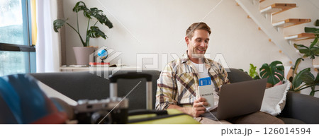 Portrait of handsome young man, has two tickets and passport, goes online on laptop, booking hotel and planning his holiday, sitting in living room 126014594