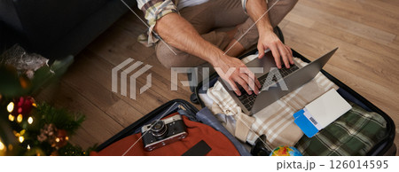 Close up shot of man, male hands using laptop on top of opened suitcase, packing for a holiday, going on vacation, prepare camera, two flight tickets for a trip abroad 126014595