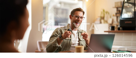 Happy young man in glasses, freelancer sitting in cafe with laptop, drinking coffee, pointing fingers at camera and smiling cheerfully 126014596