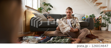 Travelling and tourism concept. Happy young man, sits on floor with luggage opened, packing suitcase for holiday, going on vacation, putting clothes inside a bag 126014647