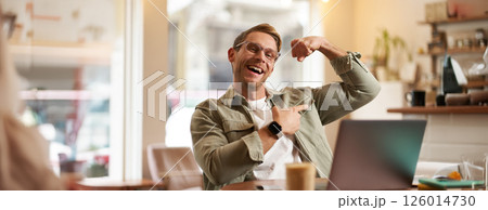 Portrait of handsome, pleased young man shows biceps, flexing muscles, sits in front of laptop in cafe, rejoicing, private coach having a video chat with his client 126014730