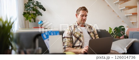 Portrait of handsome young man, has two tickets and passport, goes online on laptop, booking hotel and planning his holiday, sitting in living room 126014736