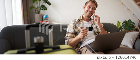 Portrait of young man with credit card, sitting and using laptop, making a phone call, booking hotel or tickets, confirming his purchase over telephone, has suitcase near him 126014740