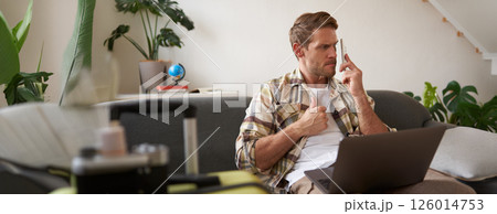 Portrait of angry man waiting on the line, trying to call someone, using smartphone and laptop, sitting near suitcase 126014753