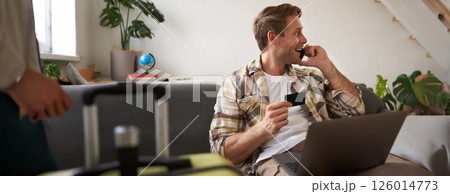Image of guy calling someone, holding credit card and using laptop, has suitcase and camera packed for holiday 126014773