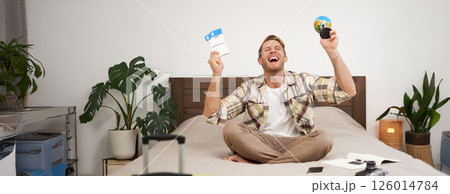 Portrait of happy guy, tourist goes on vacation abroad, holds globe and plane ticket, excited about his holiday, sits on bed with smiling, enthusiastic face expression 126014784