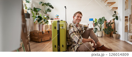 Smiling handsome guy looks at his passport and flight ticket, sits on floor with suitcase 126014809