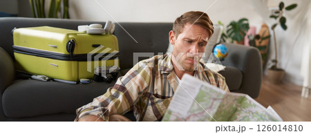 Portrait of man with concerned, puzzled face looking at map, frowning while studying the route, sitting with suitcase, going on vacation 126014810