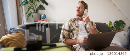Handsome smiling man with laptop, making a phone call, looking aside while waiting on the telephone, confirms booking online Handsome smiling man with laptop, making a phone call, looking aside while waiting on the telephone, confirms booking online 126014865