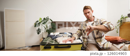 Image of young disappointed man, showing thumbs down, puts hand on his clothes inside luggage, dislike, disapprove something awful, upset with his holiday 126014903