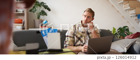 Portrait of man with concerned face, looks aside with worried expression, sits on sofa with laptop, has two tickets on plane and suitcase 126014912