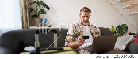 Portrait of cheerful man, going on holiday, plans a travel journey abroad, has laptop opened, sits in living room with suitcase, books a hotel Portrait of cheerful man, going on holiday, plans a travel journey abroad, has laptop opened, sits in living room with suitcase, books a hotel 126014939