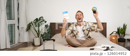 Portrait of cheerful, happy tourist, man sitting with plane tickets and a globe, packed suitcase for travelling around the world, going on vacation, excited about his holiday 126014952
