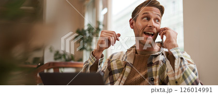 Freelance, digital nomad and workspace concept. Portrait of handsome young man with mobile phone, sitting in cafe with laptop, working on project, studying remotely, looking back outside window 126014991
