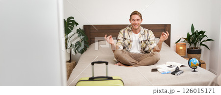 Portrait of young man meditating, packing suitcase, sitting on bed with globe, camera and plane tickets, relaxing before travelling abroad 126015171