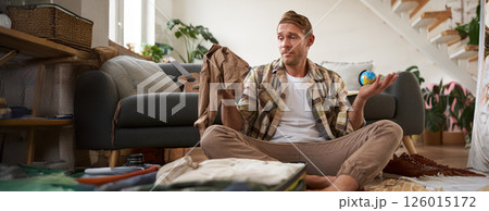 Portrait of confused man with suitcase, holding pants, shrugging shoulders and looking clueless at clothes luggage Portrait of confused man with suitcase, holding pants, shrugging shoulders and looking clueless at clothes luggage 126015172